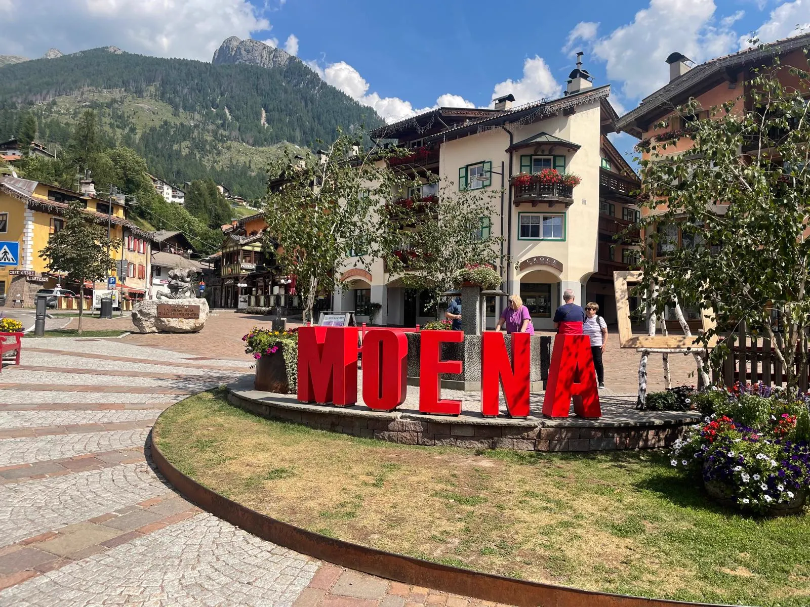 Roter MOENA-Schriftzug auf dem Dorfplatz mit Bergpanorama